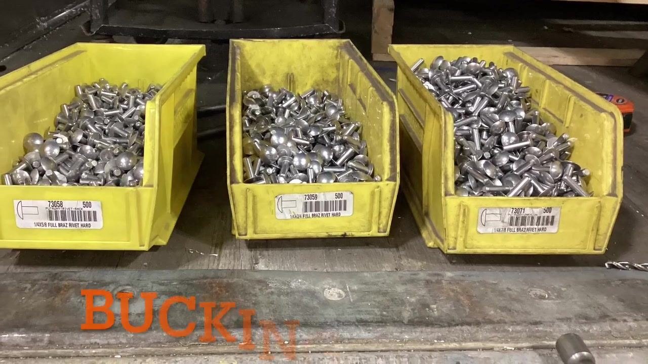 Three yellow bins labeled with different rivet sizes and filled with metal rivets. Labels read 14X5/8, 14X3/4, and 14X7/8 FULL BRAZ RIVET HARD, with the word "BUCKIN" overlaid in orange text.