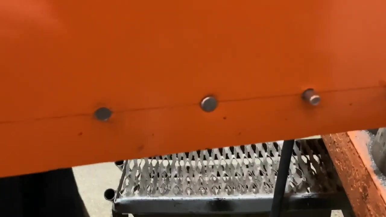 Gloved hands holding a tool near a rivet and vise, with an orange workbench and metal panel visible.