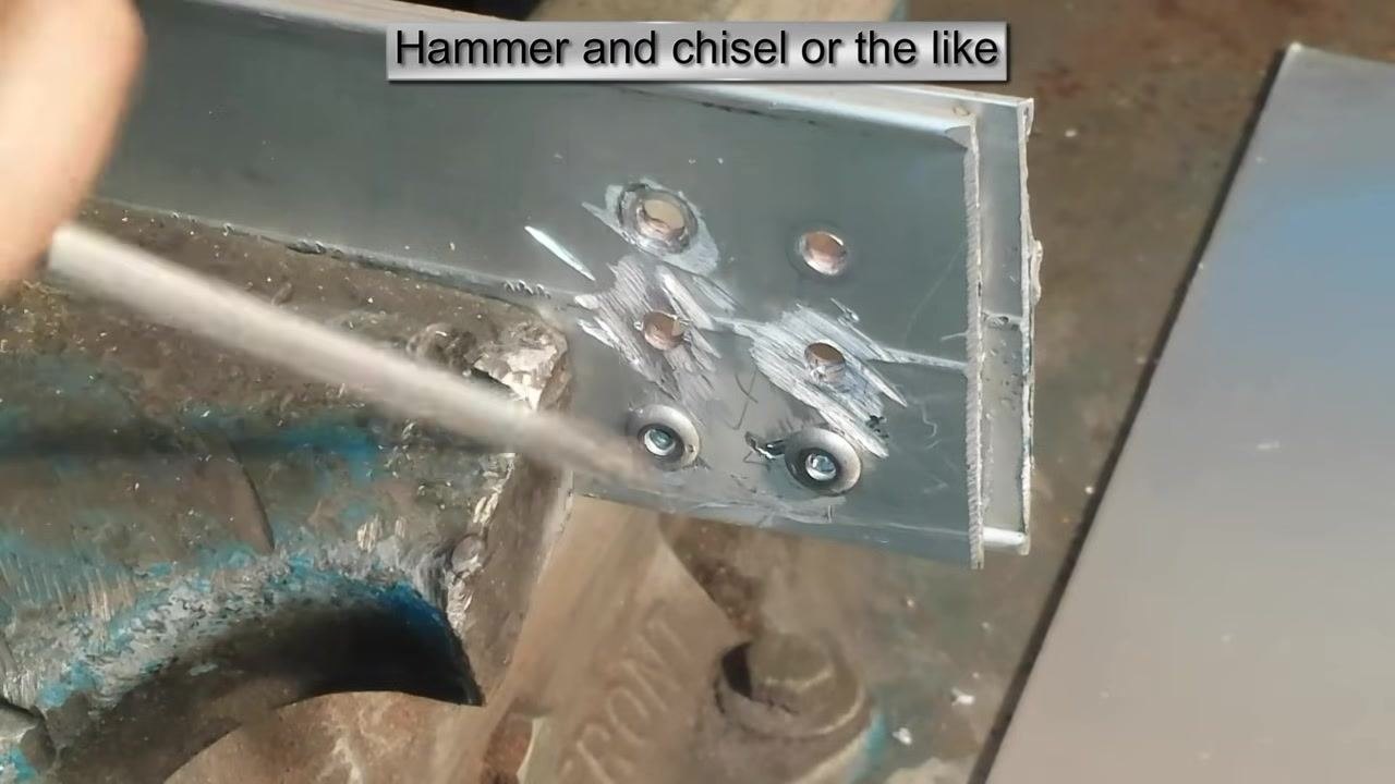 A chisel or screwdriver positioned at the edge of a rivet head on a metal L-bracket clamped in a vise, with tool marks visible from previous removals. On-screen text: "Hammer and chisel or the like."