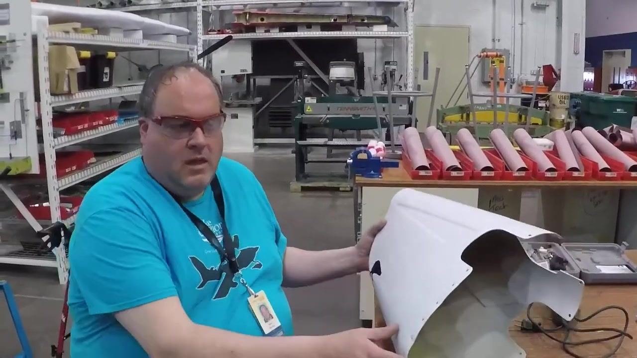 A person in safety glasses and a blue Lake Superior College t-shirt sits at a workbench in an aviation workshop, holding a white Cessna 182 tail cone with visible damage near the tip. Organized shelves, tools, and composite material storage are visible in the background.