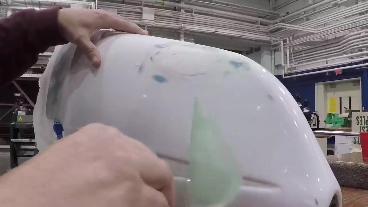 A hand touches a greenish fiberglass patch applied to a white curved object on a workbench, with various workshop items visible in the background.