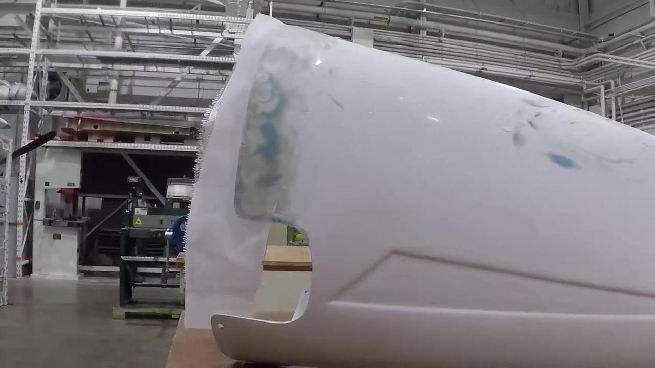 A white curved object shows visible blue and beige repair marks, resting on a workbench in a workshop, with a red box of materials and a cardboard box labeled "WORLD'S FINEST APPLES" in the background.