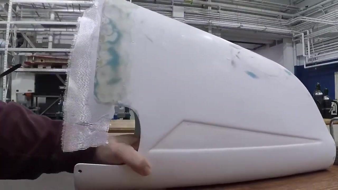A hand holds a white curved object with a freshly applied fiberglass patch that extends beyond the edge, in an industrial workshop with shelving and overhead pipes.