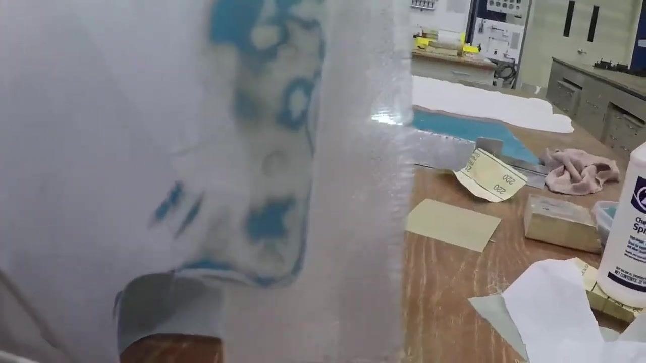 A close-up view of a hand inspecting a fiberglass patch on a white curved object; the patch is semi-transparent, and the workbench contains sanding blocks, a bottle labeled "Zep Chemical Spray," and other supplies.