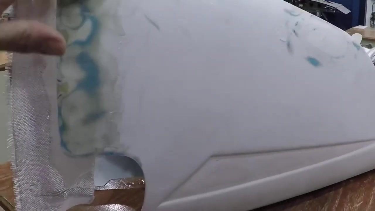 A close-up view of a fiberglass patch on a white curved object shows a workbench in the background containing sanding blocks, a bottle labeled "Zep Chemical Spray," and other supplies.