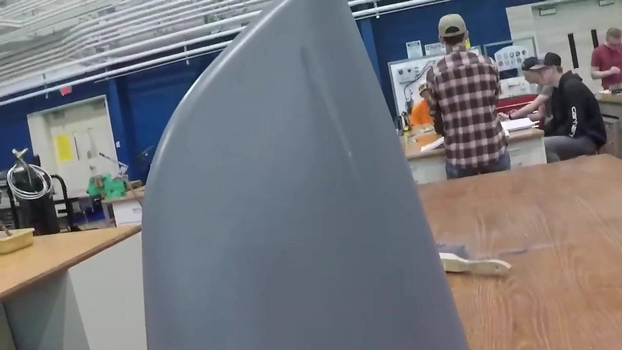 A close-up view of a hand holding a large, gray-primed fiberglass part in a workshop; workbenches, tools, and several people working on projects are visible in the background.