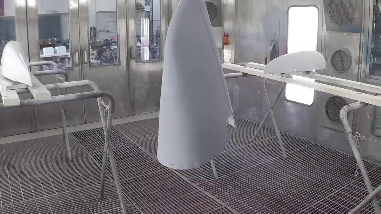 A gray-primed fiberglass part is held up in a workshop, showing a smooth, even surface; workbenches, a broom, and people working are visible in the background.