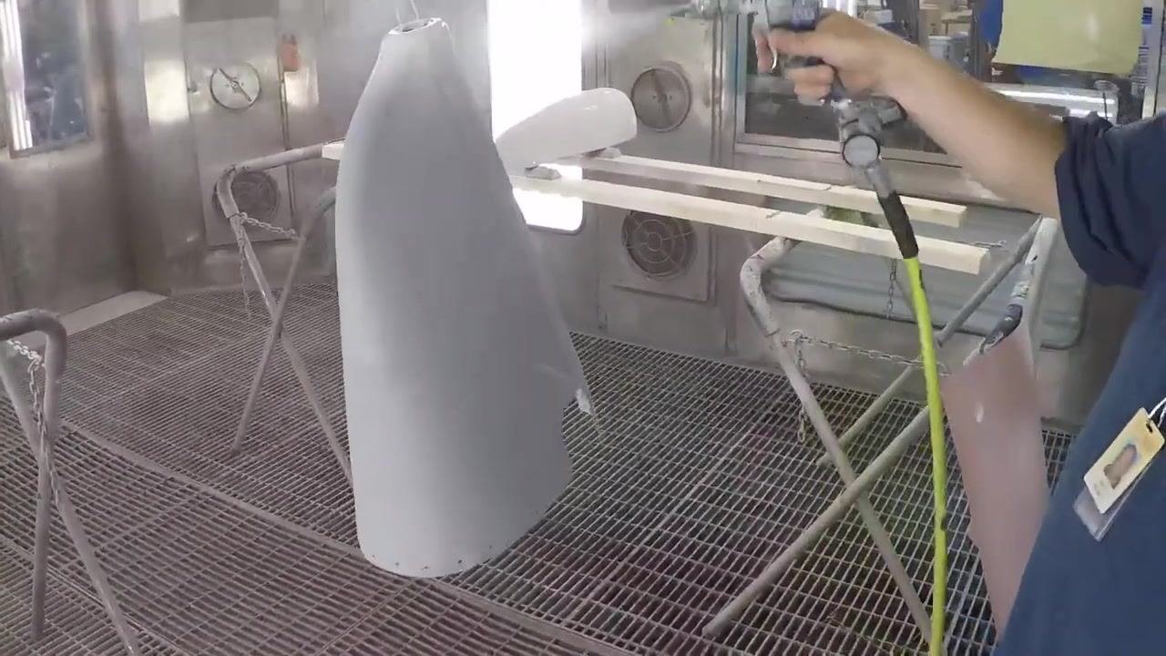 One person holds a large, gray-primed fiberglass part while another uses a spray gun to apply paint; both are in a paint booth, and the holder is wearing a respirator.