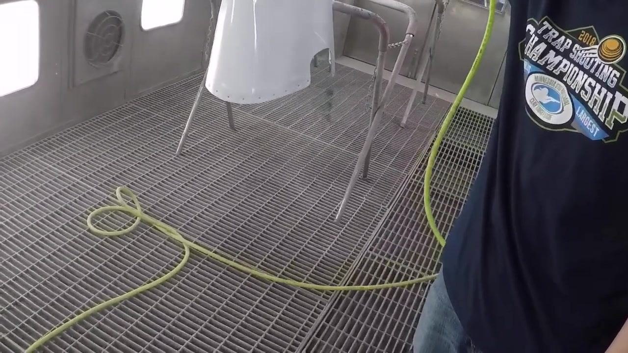 A person holds a painted fiberglass part in a spray booth, with a yellow air hose on the floor and spray equipment visible; the booth has metal grating and bright lighting.