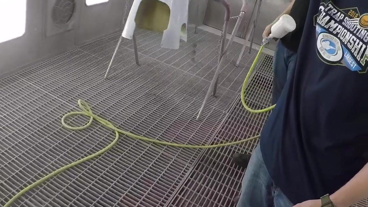 A freshly painted, glossy white fiberglass part is suspended in a professional spray booth; a person wearing a respirator and a "Trap Shooting Championship" shirt holds the part up for inspection, with bright lighting and clean surroundings.