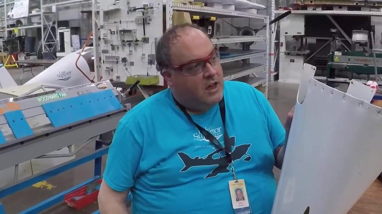 A person in safety glasses and a blue Lake Superior College t-shirt holds the Cessna 182 tail cone, with the workshop environment, including tools and aircraft parts, visible in the background.