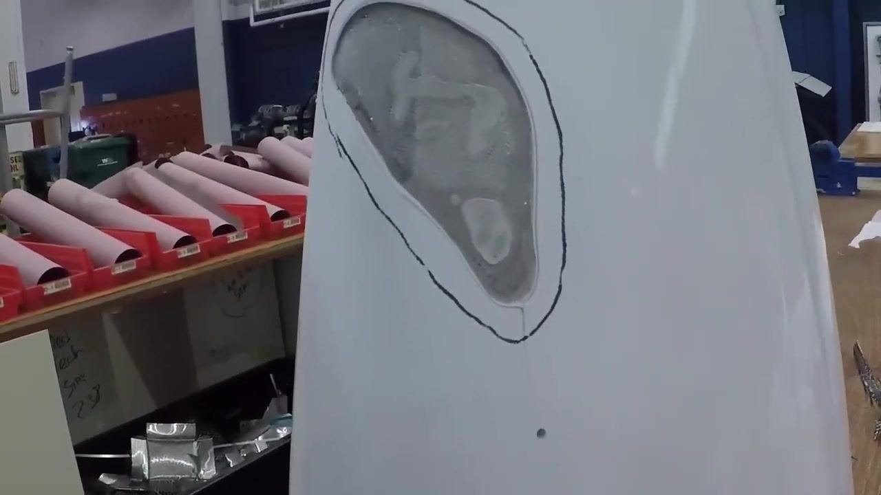 The tail cone shows fiberglass laid over the repair area, with the pen outline still visible and the surface prepared for curing; the workshop background includes composite rolls and tools.