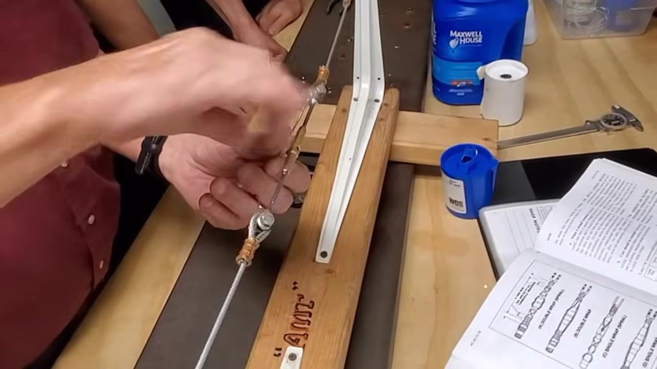A person holds a cable assembly mounted on a wooden jig labeled "TUG." The assembly includes threaded brass turnbuckles, cable ends, and clevis fittings, with tools and a manual visible on the workbench.