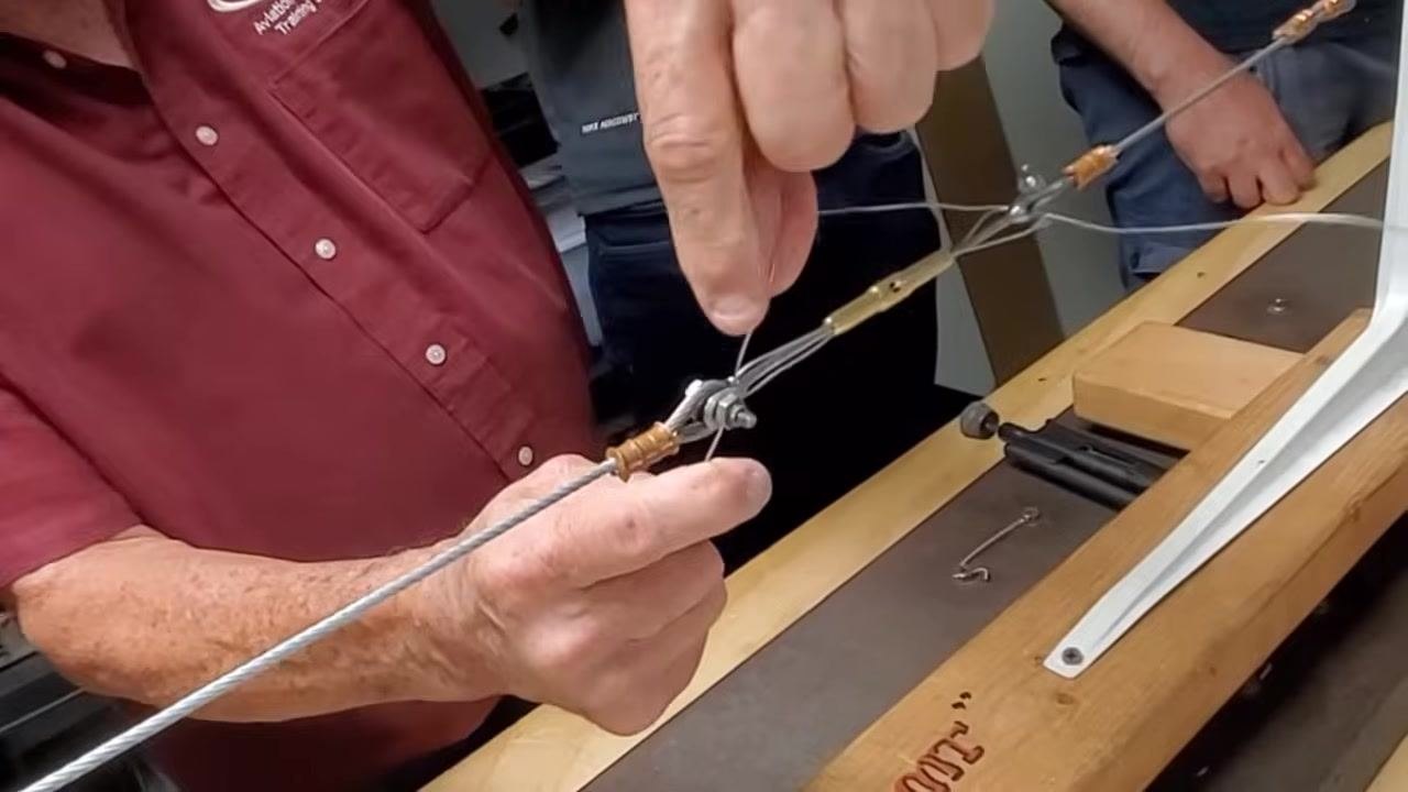 A person folds one end of the safety wire over the horn of a turnbuckle mounted in a wooden jig labeled "TUG," with tools and another person observing on the workbench. The wire is bent sharply and pressed against the horn.