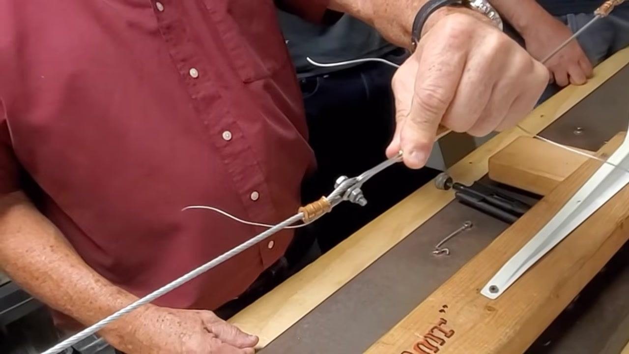 A person folds and starts twisting a safety wire around a cable clamp on a steel cable assembly by hand, on a wooden workbench with tools nearby.