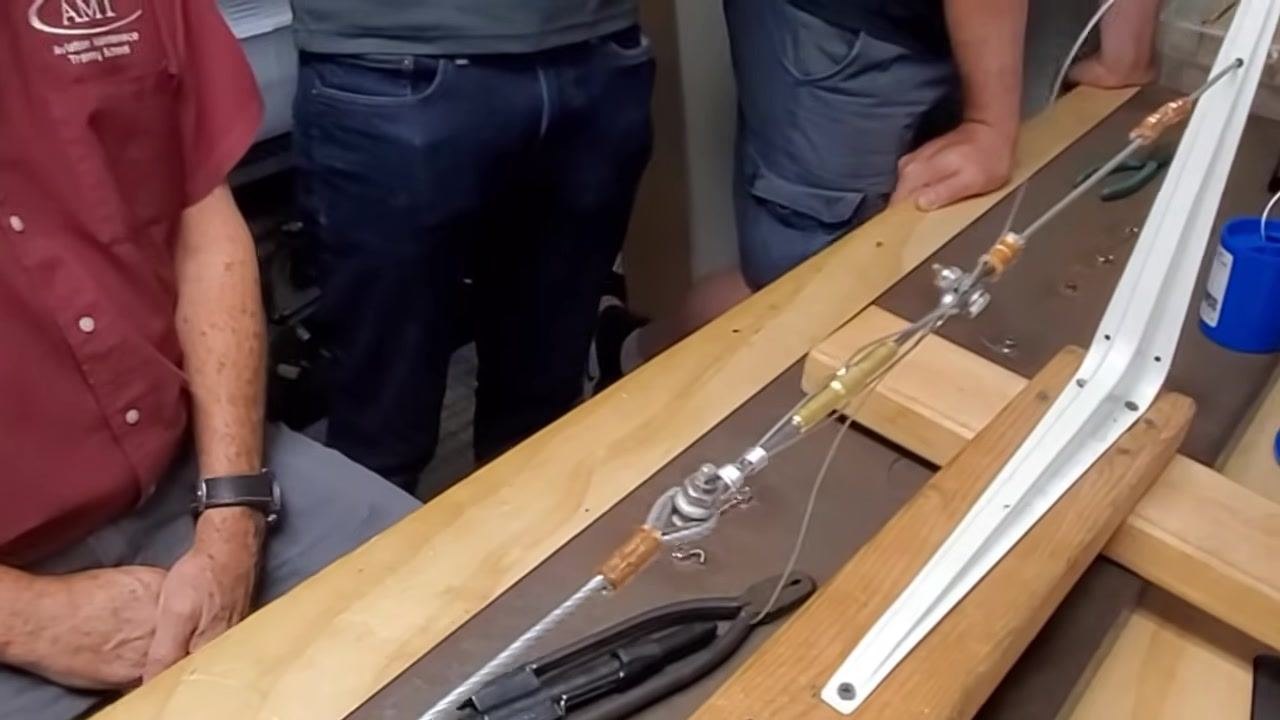 A close-up view of a hand checking the tightness and direction of the safety wire twists on the cable assembly.