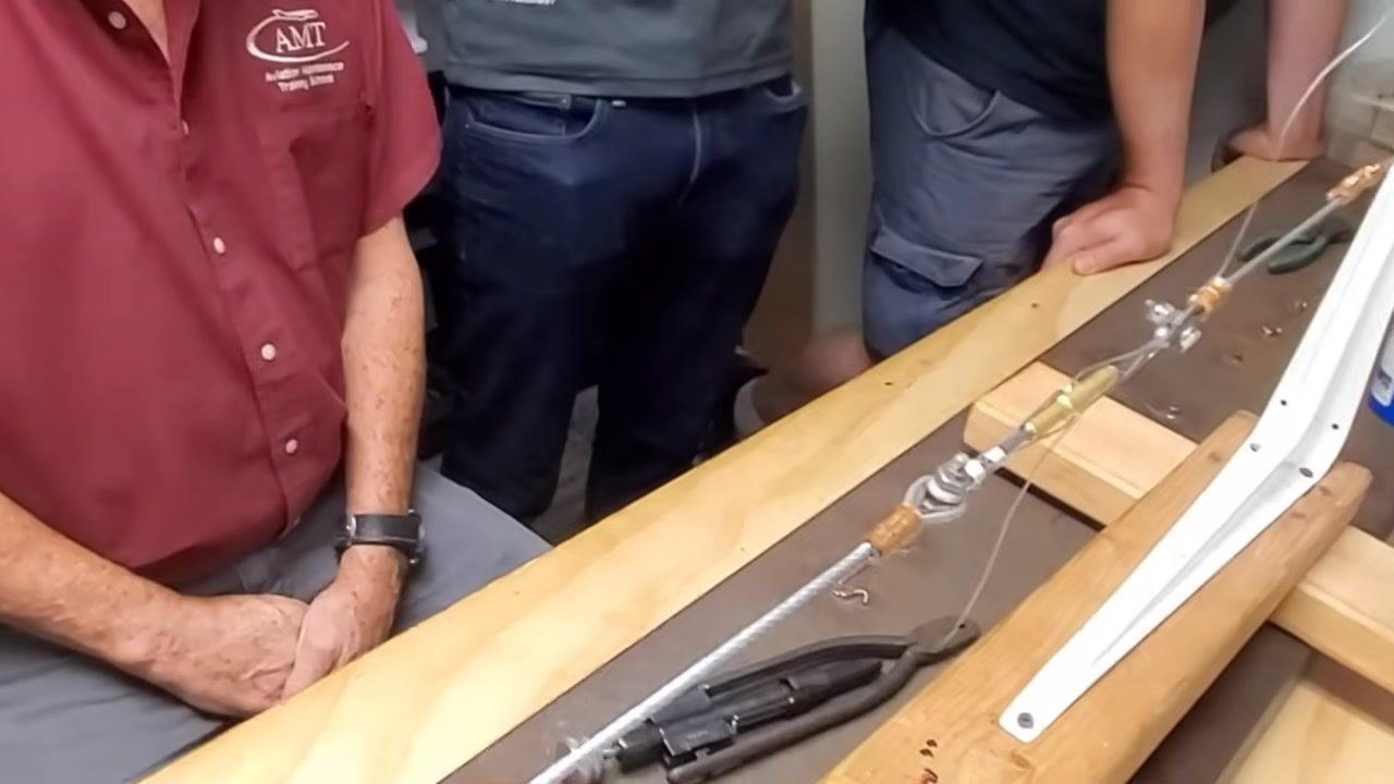 A group of people gather around the workbench, one seated and others standing, observing the completed cable assembly and preparing for the next person to try the procedure.
