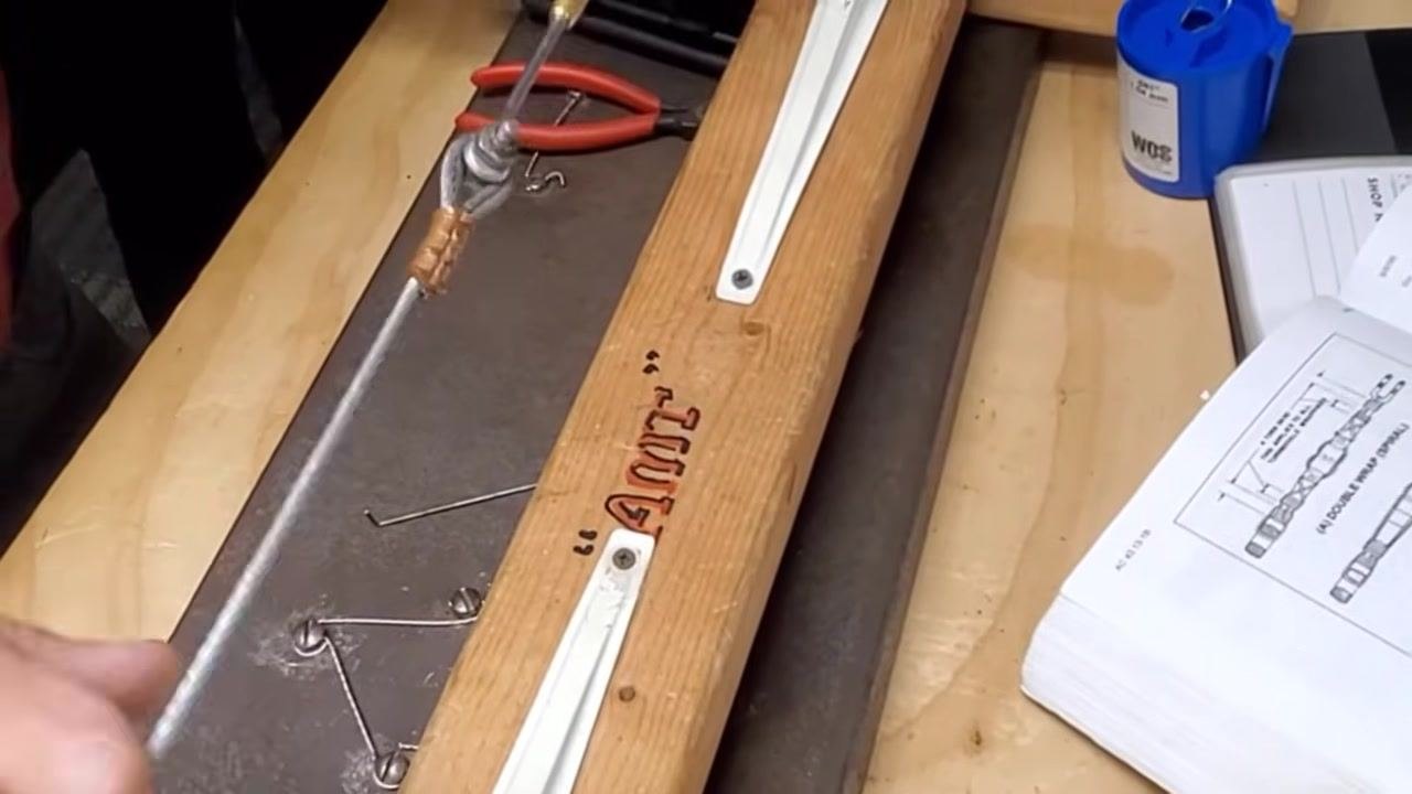 The completed cable assembly is mounted on the wooden jig, with tools and a manual nearby. The threads are evenly engaged and the assembly appears secure.