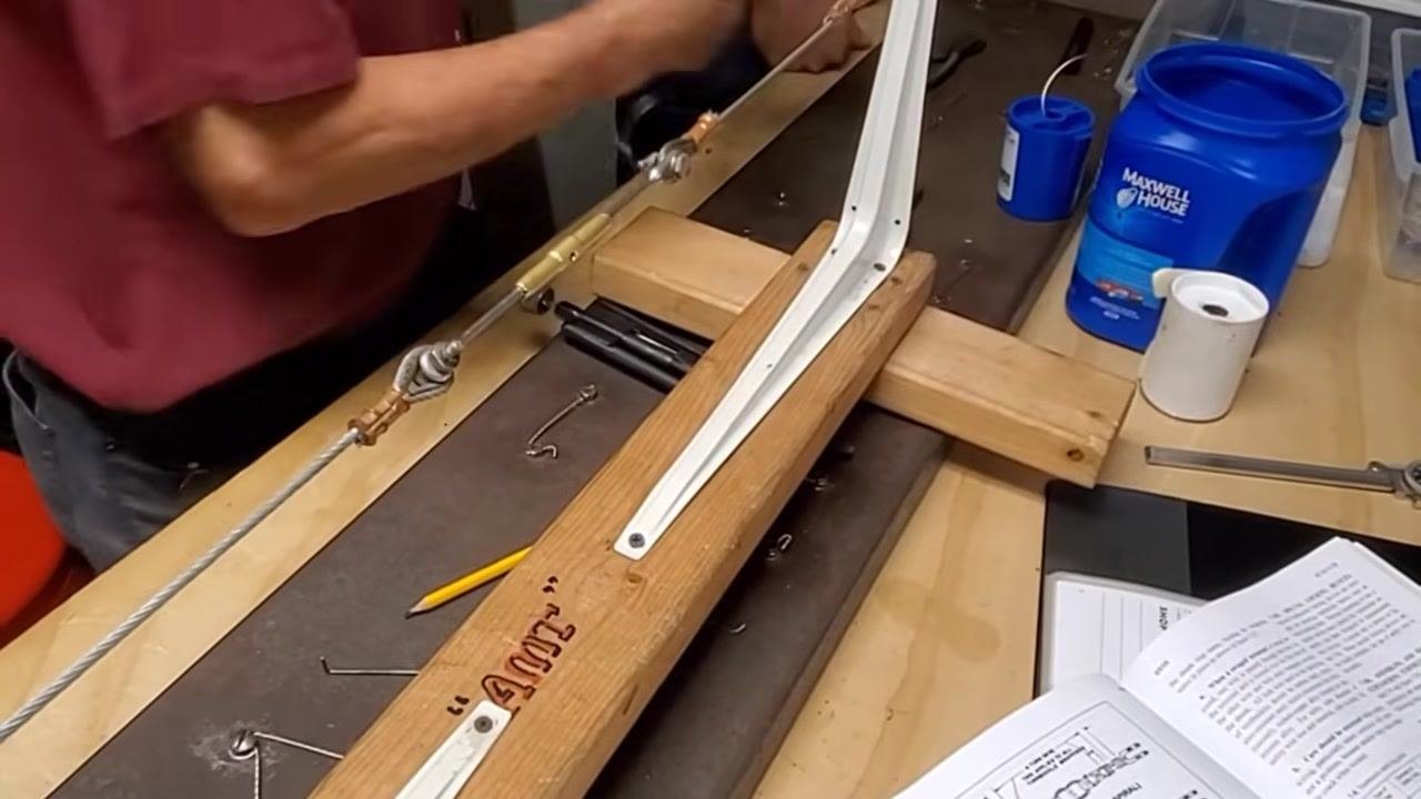 A person holds a piece of safety wire, preparing to insert and bend it through the turnbuckle assembly, with tools, the cable assembly, and the open manual on the workbench.