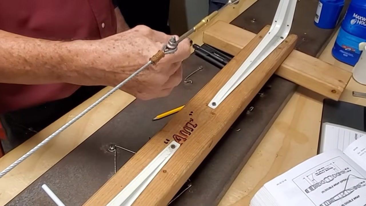 A person threads a piece of safety wire through the horn of a turnbuckle on a cable assembly mounted in a wooden jig labeled "TUG," with tools and an open manual on the workbench.