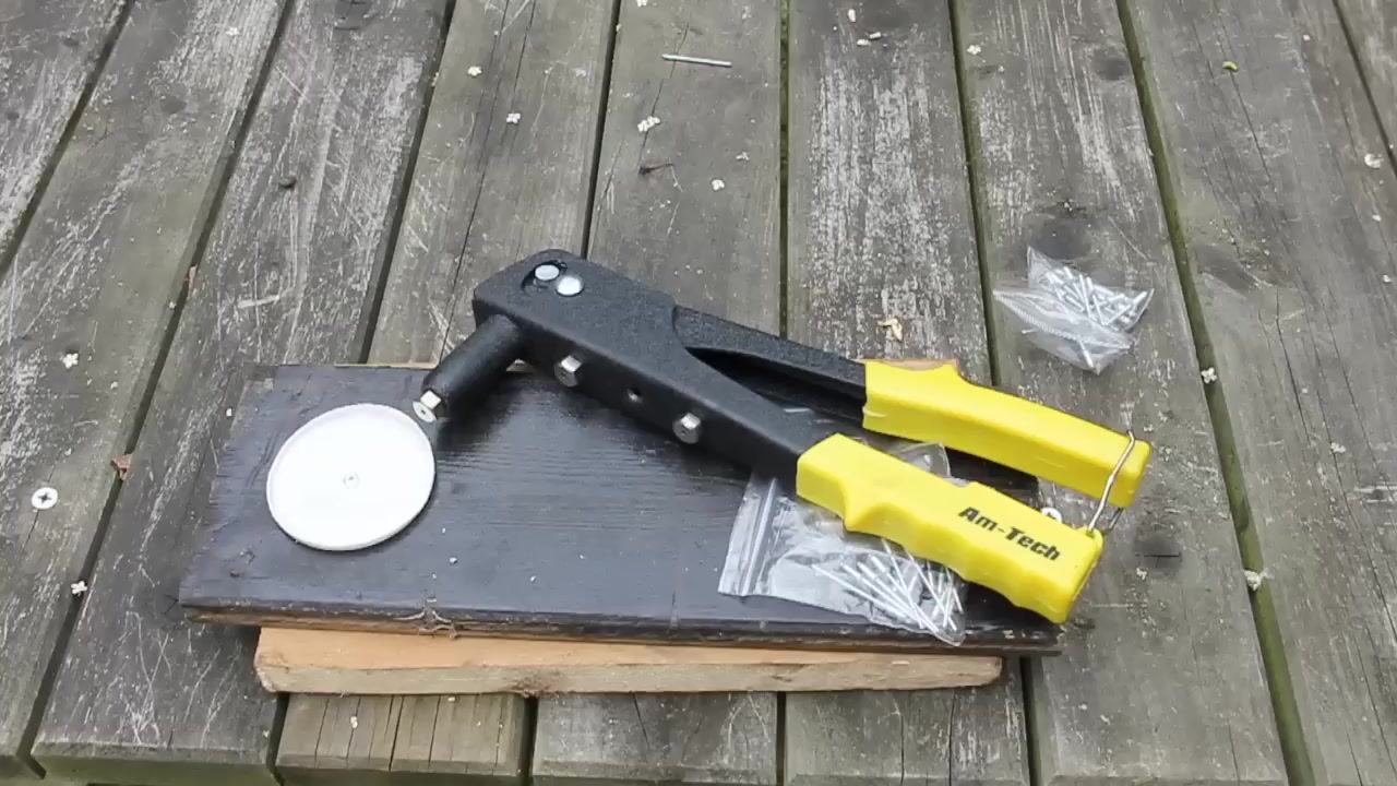 The user squeezing the handles of the yellow-handled pop riveter positioned over the plastic and wood materials, with a bag of rivets visible nearby on the workbench.