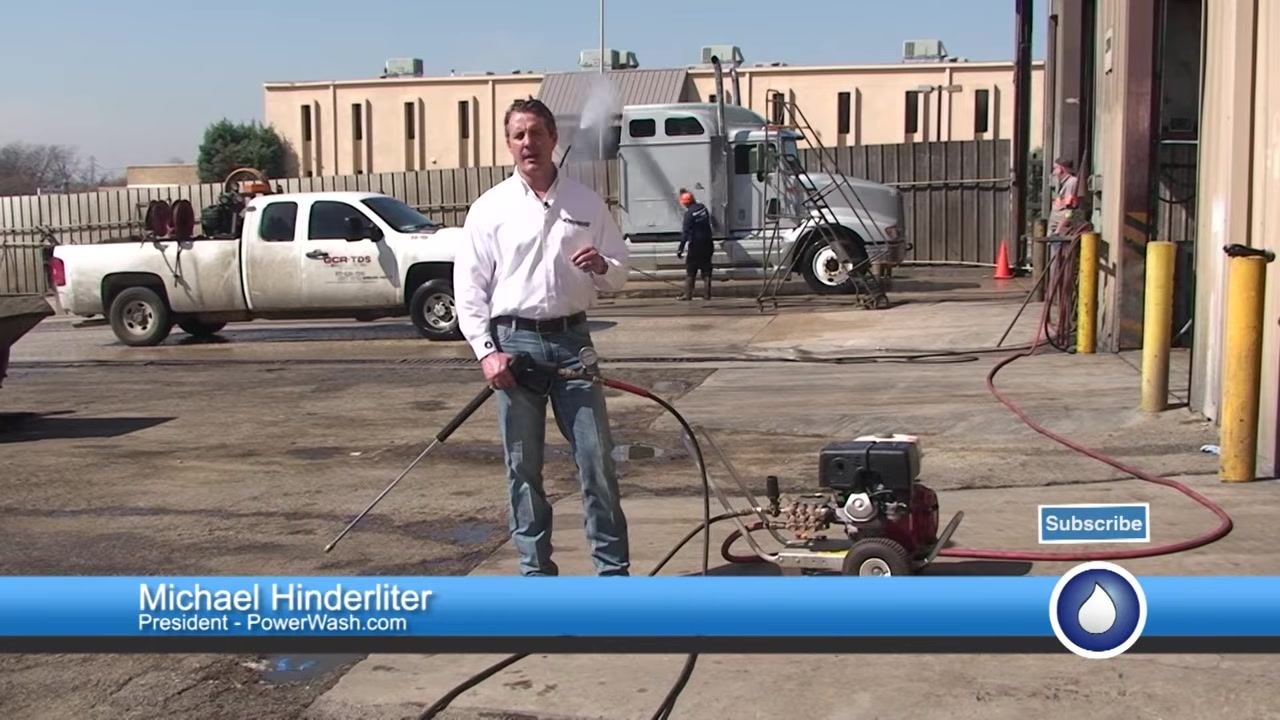 A person in a PowerWash.com shirt stands outdoors holding a pressure washer gun with a test gauge attached, with trucks and equipment in the background.