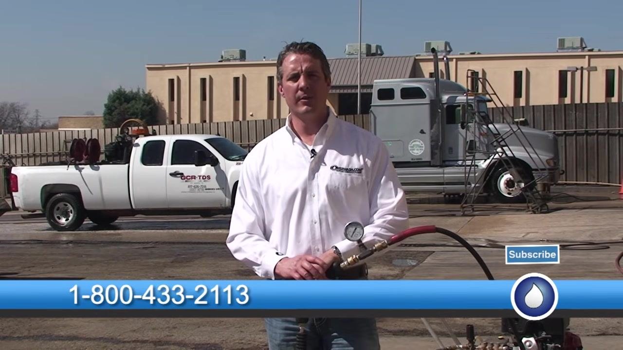 Presenter standing outdoors in a work area, holding a spray gun with a pressure gauge. Company vehicles (white pickup truck labeled GCR-TDS and a large gray semi-truck) are visible in the background. A blue banner at the bottom displays the phone number 1-800-433-2113, with a "Subscribe" button and the PowerWash.com logo.