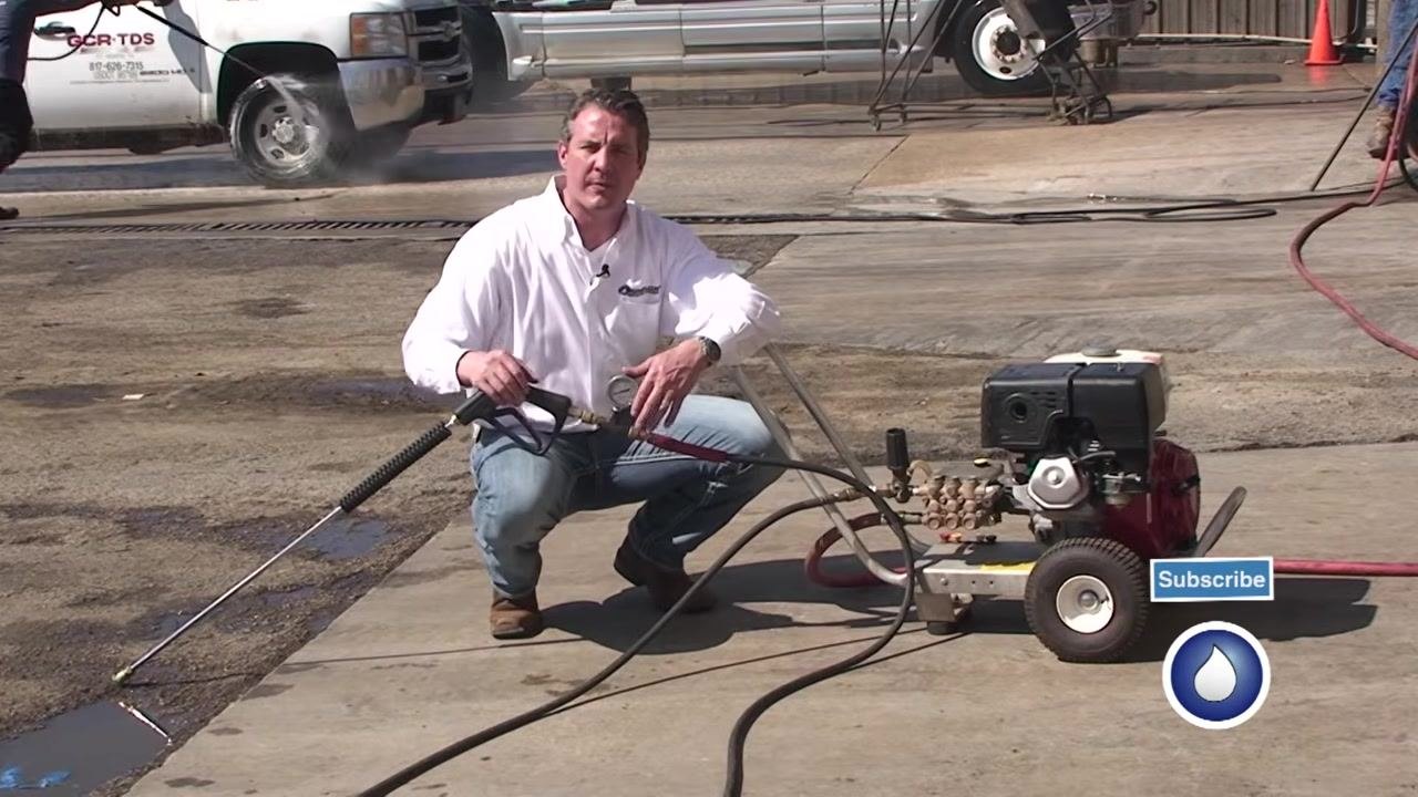 The spray gun and test gauge are held up in the outdoor work environment with the pressure washer visible.