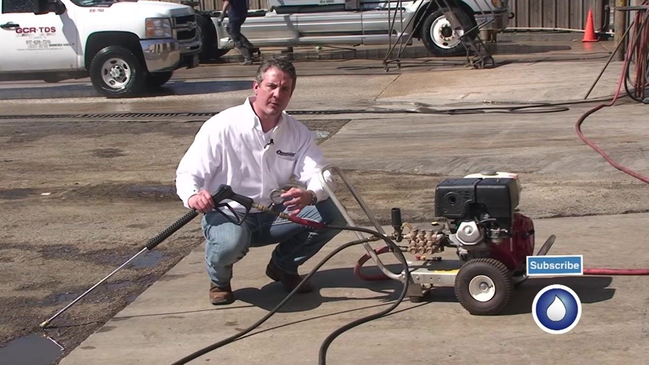 The spray gun with test gauge attached is held while preparing to pull the trigger, with the pressure washer running in the background.