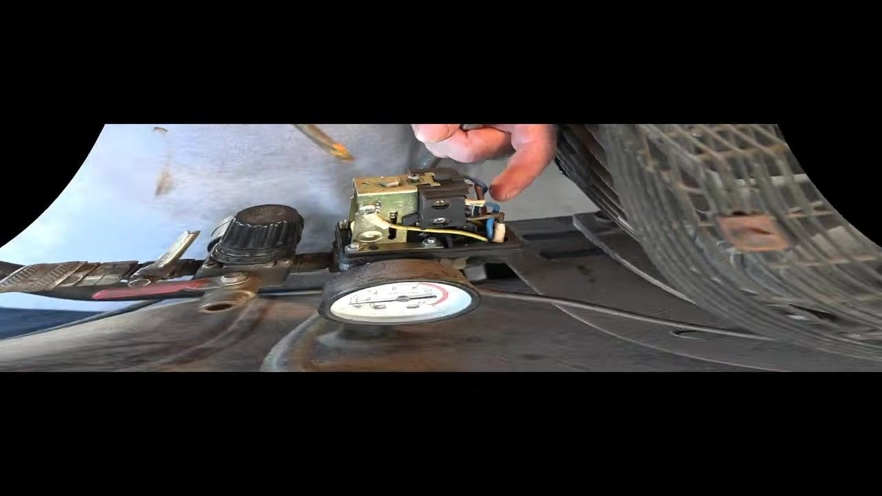 Hand using a screwdriver to adjust the top screw on the pressure switch, with the pressure gauge and wiring visible