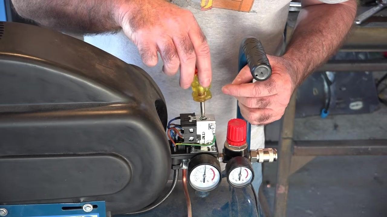 Phillips screwdriver pointing at the adjustment screw on top of the pressure switch, with dual gauges and the red regulator knob visible