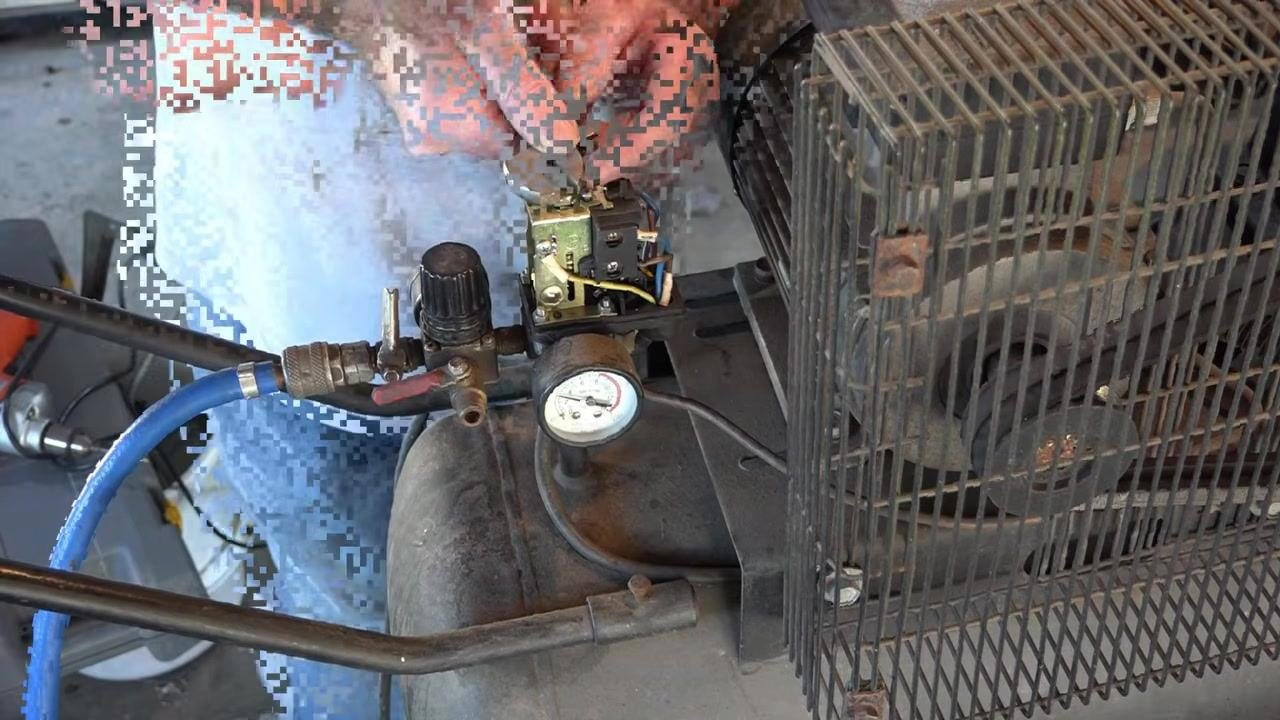 Person standing in front of the compressor holding a tool, with the pressure switch exposed, the compressor unplugged, and the blue air hose and pressure gauge visible