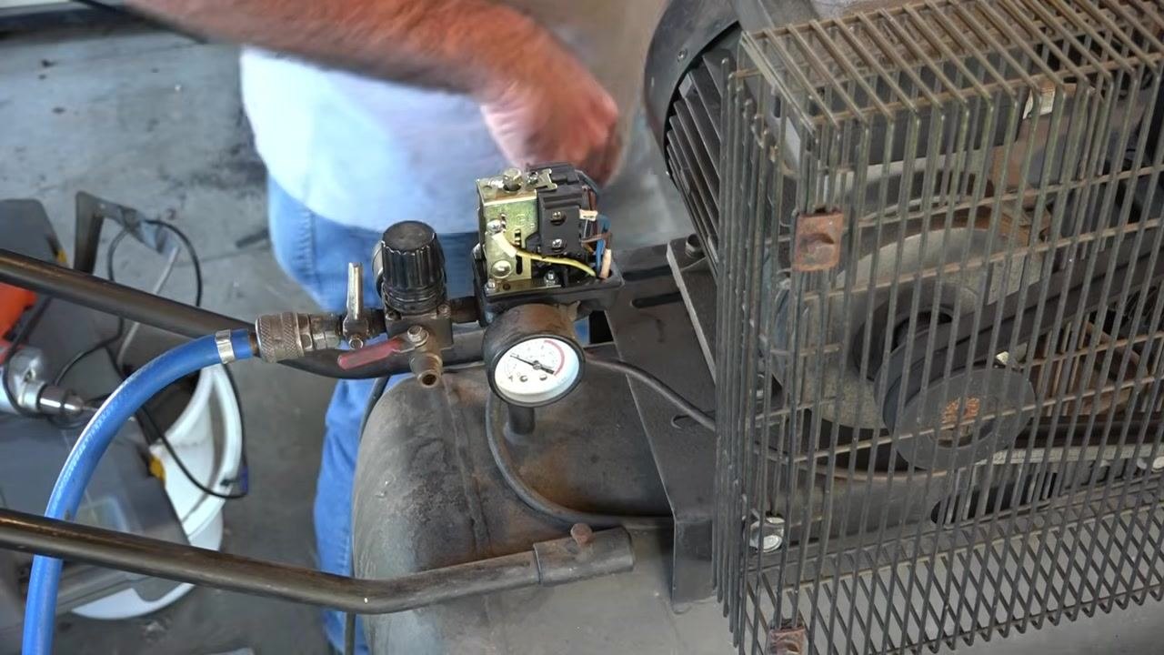 Air compressor with an exposed pressure switch, pressure gauge, regulator, and blue air hose in a workshop with tools in the background