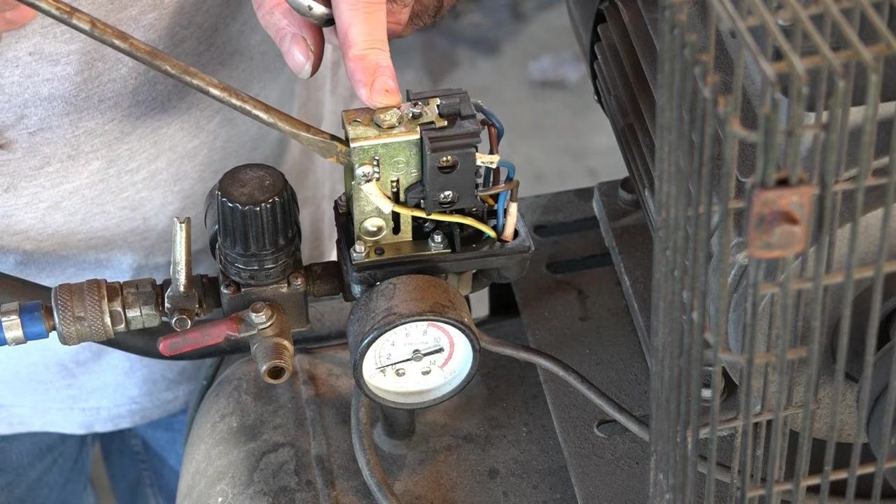 Close-up of the pressure switch with the cover removed, showing a hand pointing at the adjustment screw and spring, with gauge, wiring, and regulator visible
