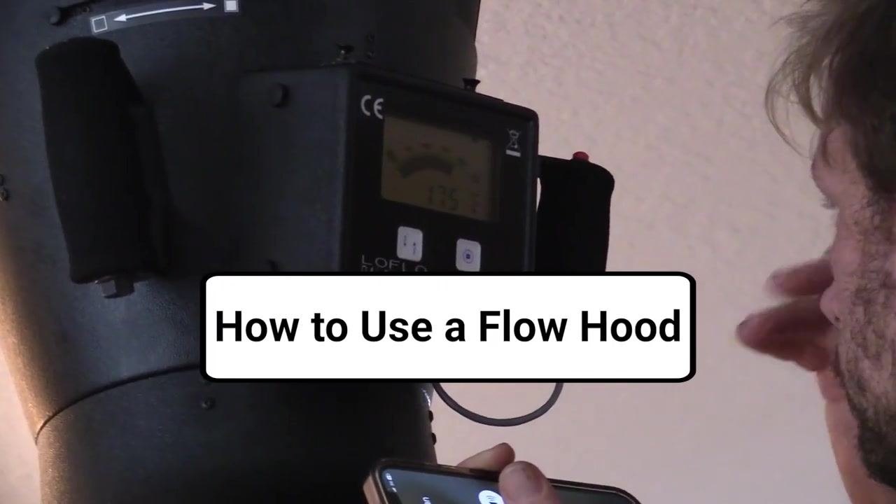 A person holds a flow hood up to a duct in a residential hallway, preparing to take an airflow measurement. The device display is visible.