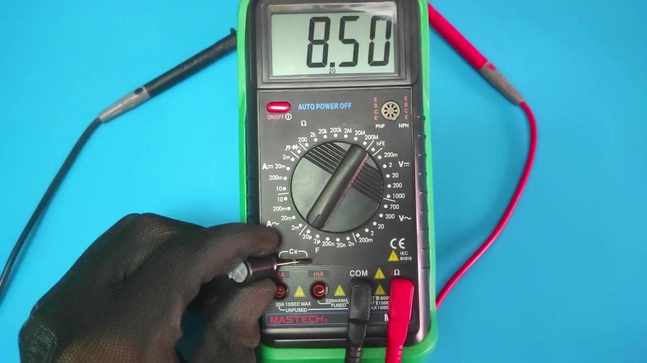Two gloved hands connecting a 9V battery to a capacitor using red and black jumper wires on a blue surface.