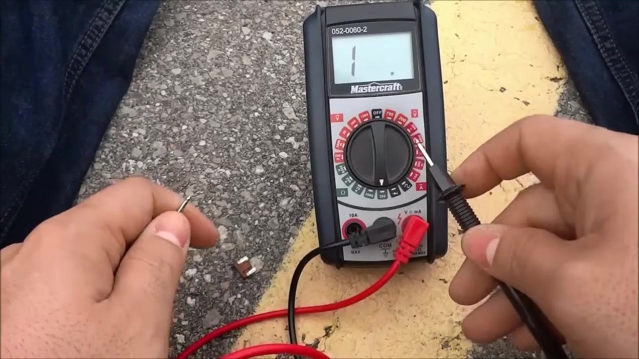 Hands hold a car fuse while touching each terminal with the red and black test leads; the display shows "1", indicating no continuity at this moment.