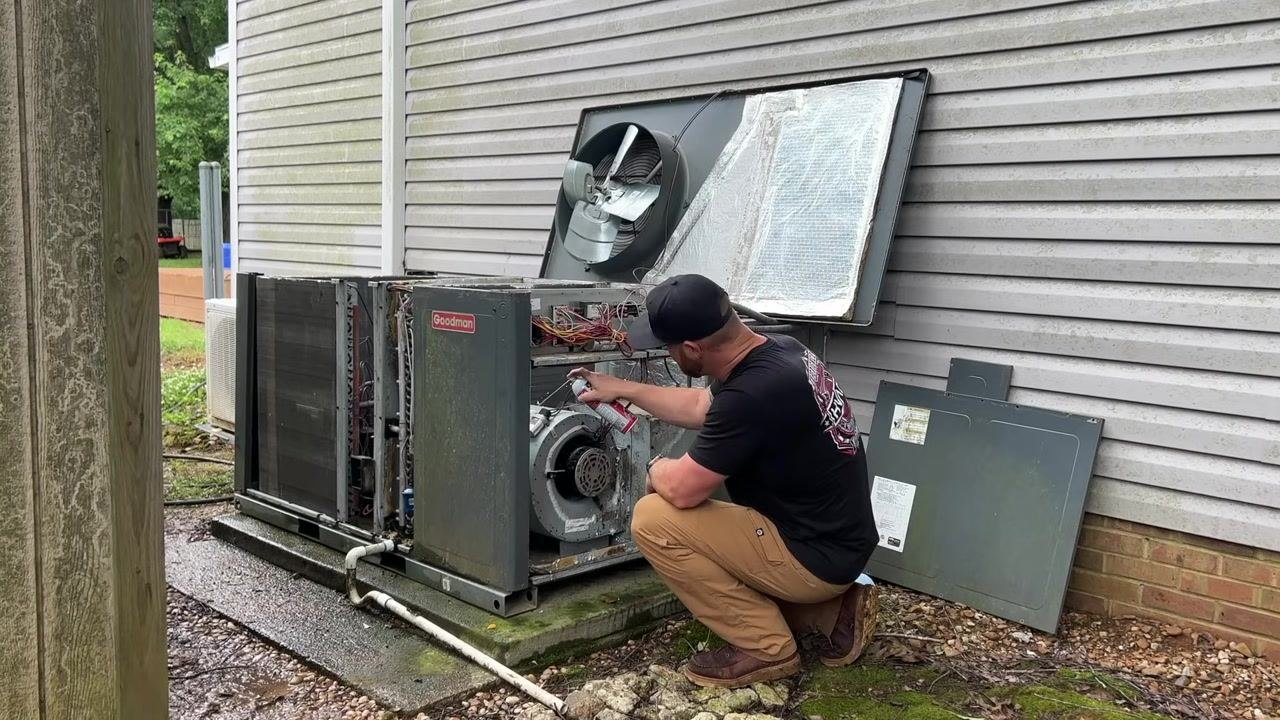 A technician kneels and sprays cleaner or lubricant onto the blower motor inside the open HVAC unit.