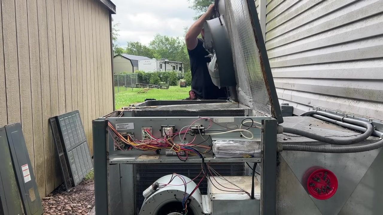 A technician lifts the fan assembly back onto the HVAC unit, with wiring and internal components visible.