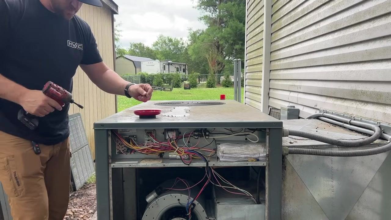 A technician holds a power drill and a screw, preparing to reattach the top panel of an open HVAC unit. A magnetic tray with screws sits on top of the unit, with internal wiring visible in an outdoor setting.