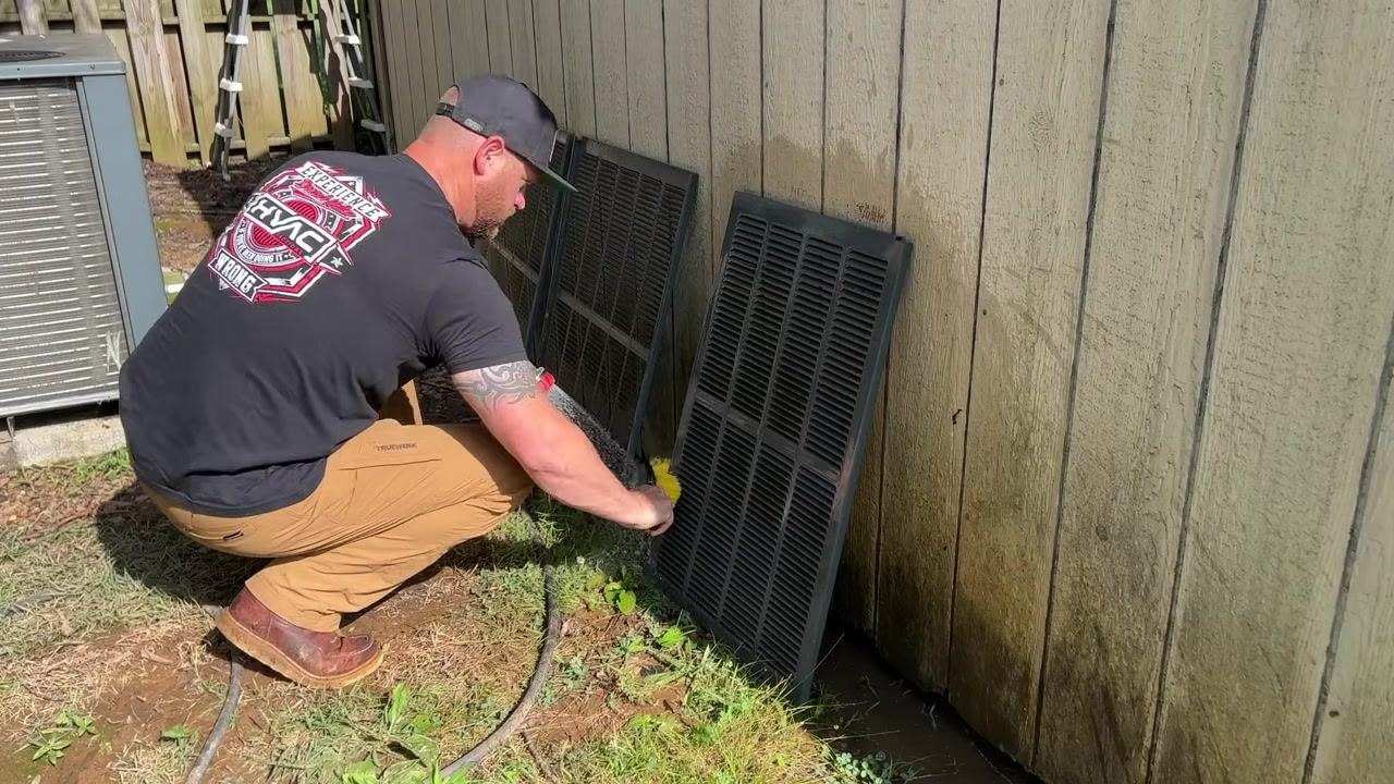 A technician kneels and scrubs HVAC grilles with a brush and hose; the grilles are propped against a wall in an outdoor setting.