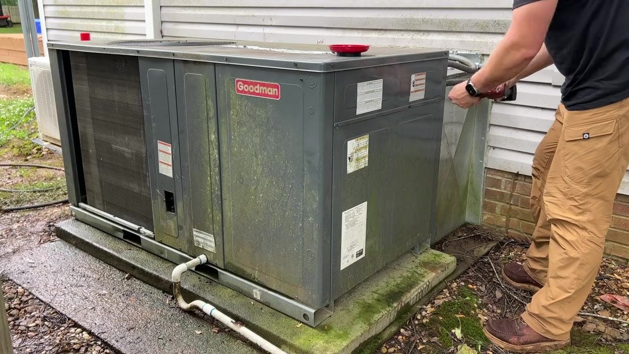A technician finishes securing the HVAC panel with a power tool; the unit sits outdoors next to a building, with visible piping and labels.