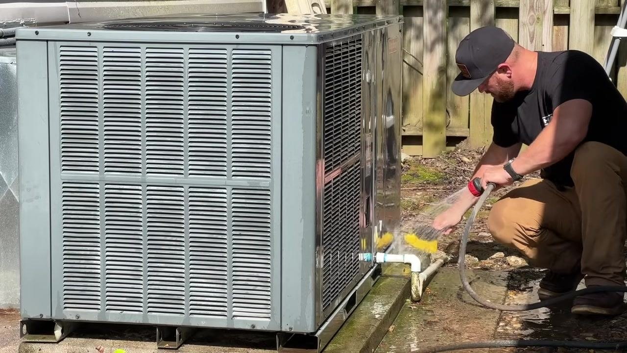 A technician uses a hose and brush to wash the exterior shroud and piping of the HVAC unit outdoors, with water and cleaning tools visible.