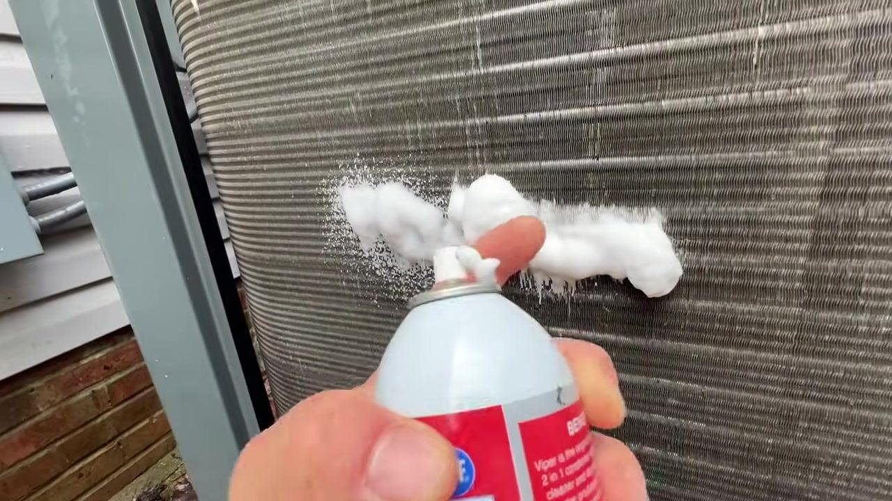 A hand holds a can of foaming coil cleaner, spraying a thick layer of foam onto the metal fins of an outdoor condenser coil. The foam is visibly expanding on the surface. The setting is outdoors, next to a building.