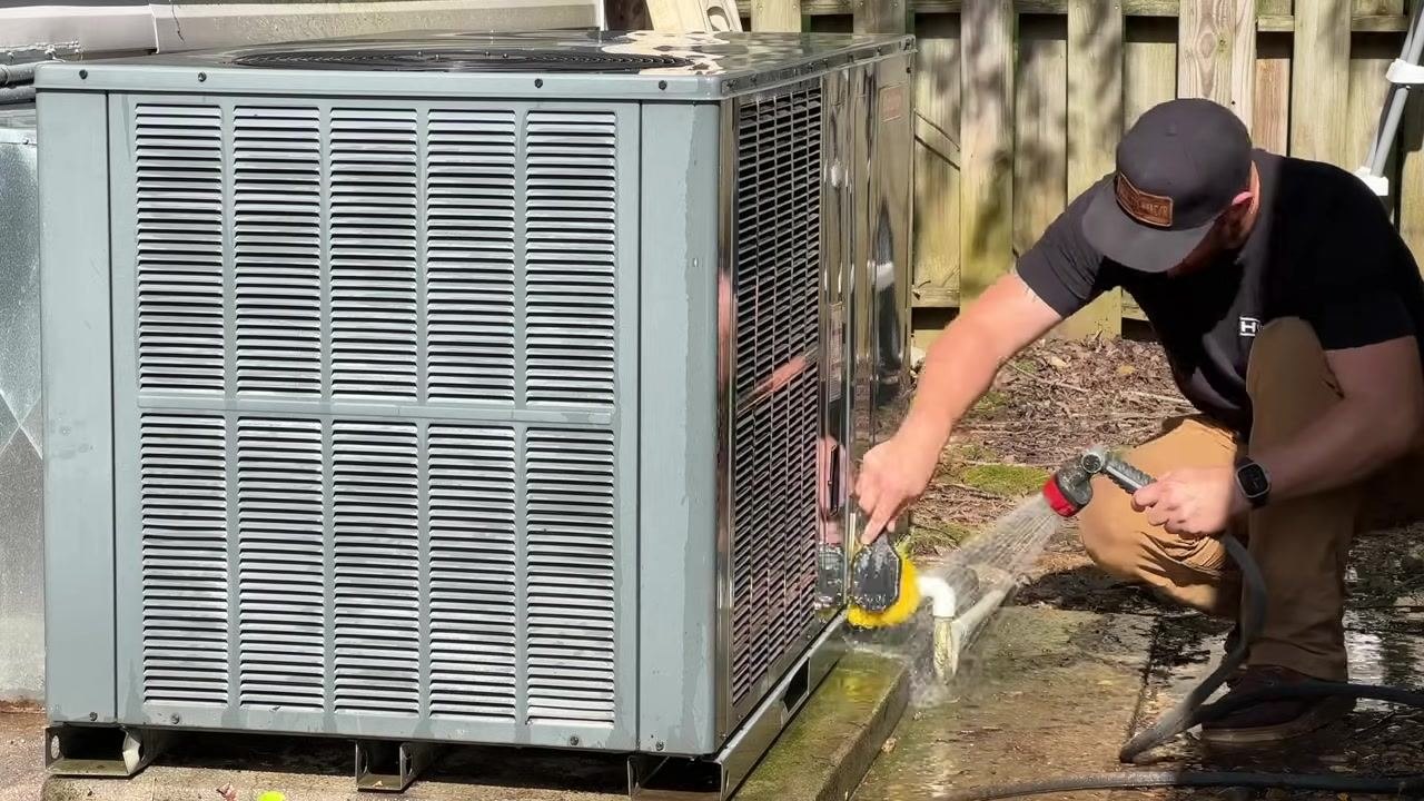 A technician scrubs and rinses the lower section of the HVAC unit with a hose and brush, focusing on thorough cleaning.