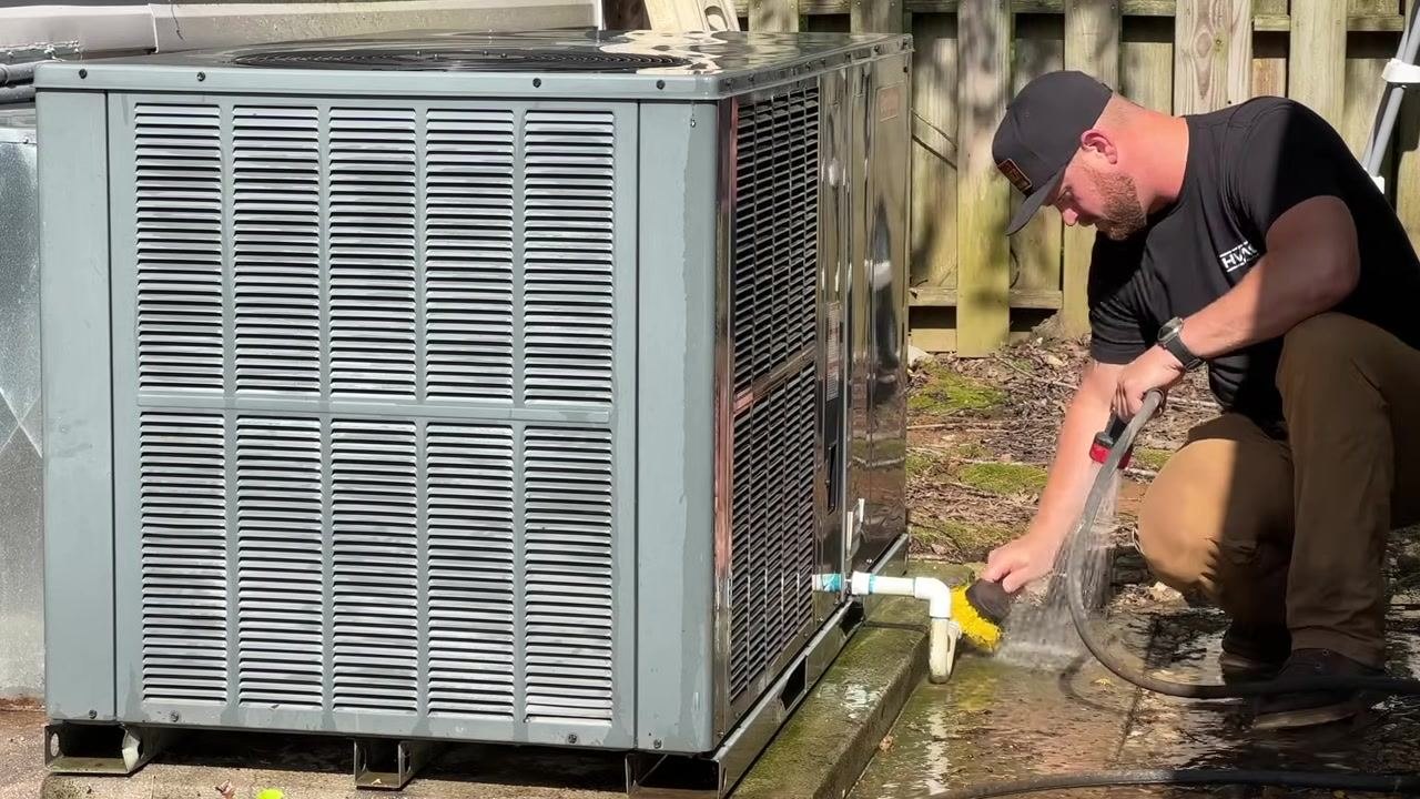 A technician rinses and scrubs the drain line and base of the HVAC unit, ensuring all components are clean.