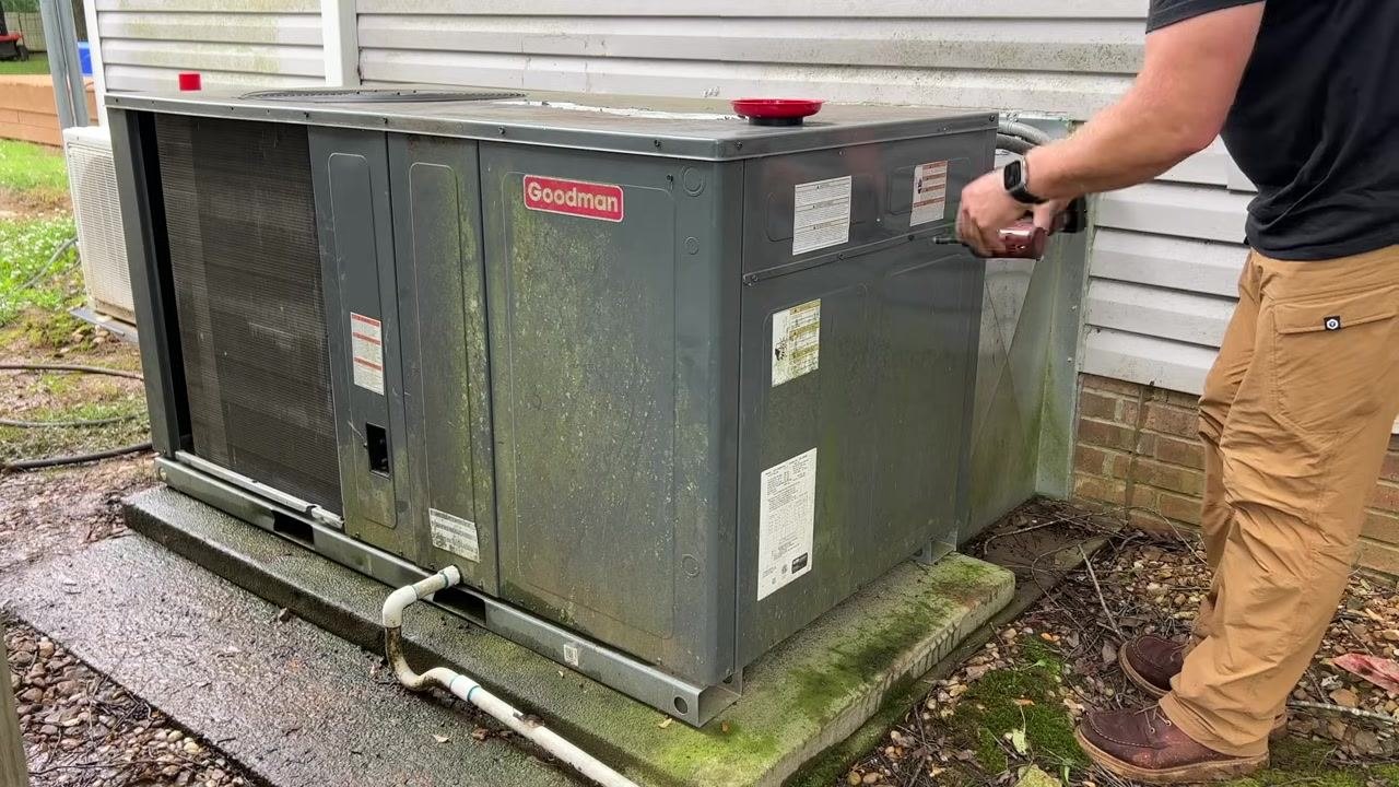 A technician uses a power tool to secure the access panel on a Goodman HVAC unit mounted on a concrete pad, with visible labels and piping.