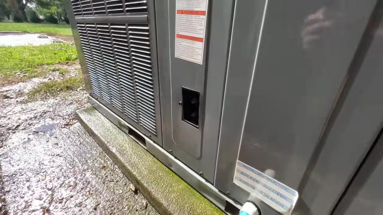 A close-up of the HVAC unit's service port area shows an open port on the side panel, warning labels, and a clean concrete pad.