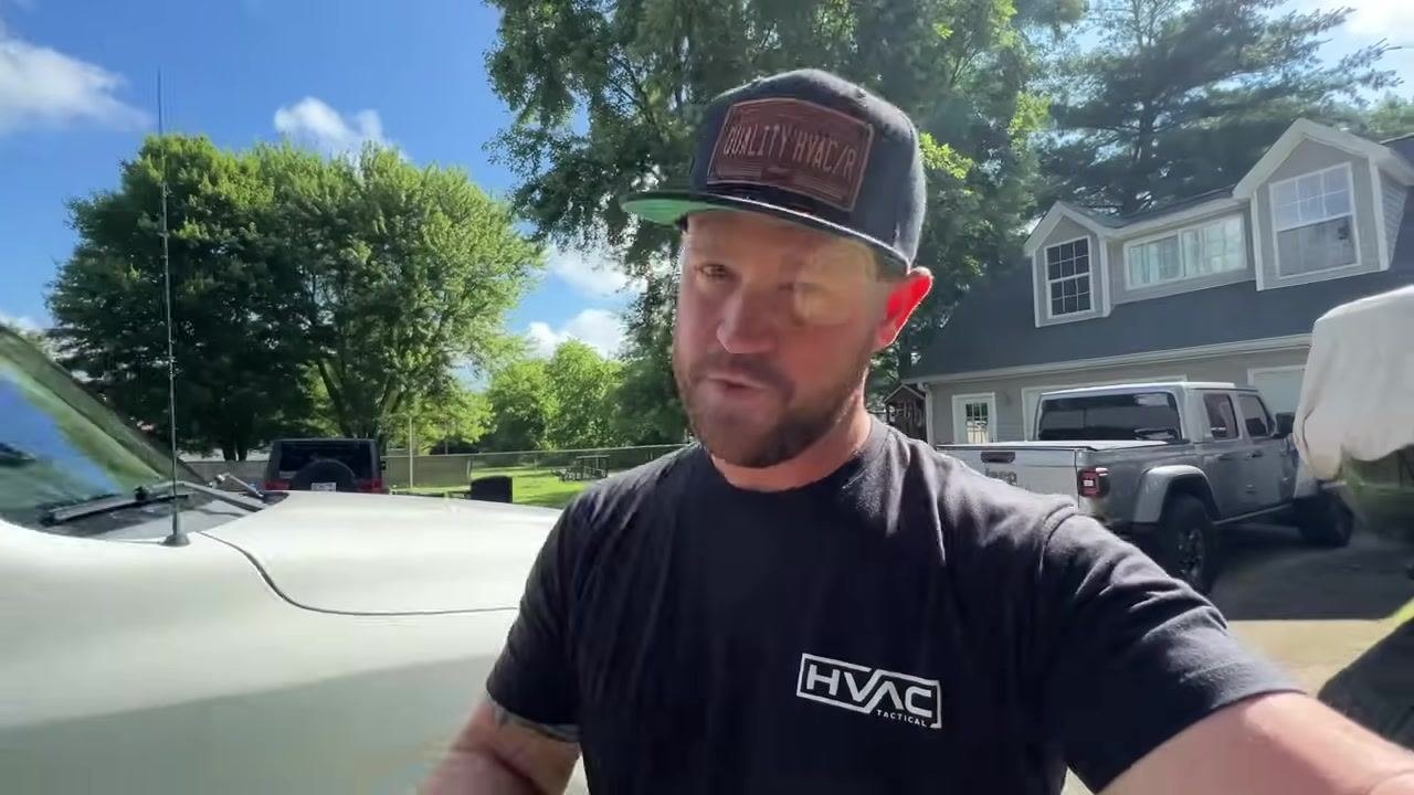 Technician in a black "HVAC Tactical" shirt and "QUALITY HVAC/R" cap, standing in a driveway next to a white vehicle, gesturing toward the camera with a house, vehicles, and trees in the background.