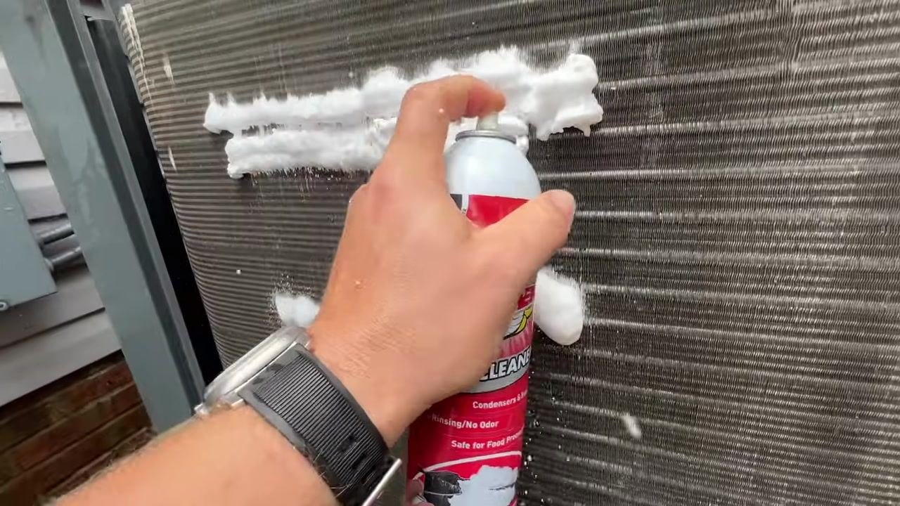 A hand sprays foam cleaner onto the condenser coil, covering a larger area. The foam forms thick lines across the coil fins.